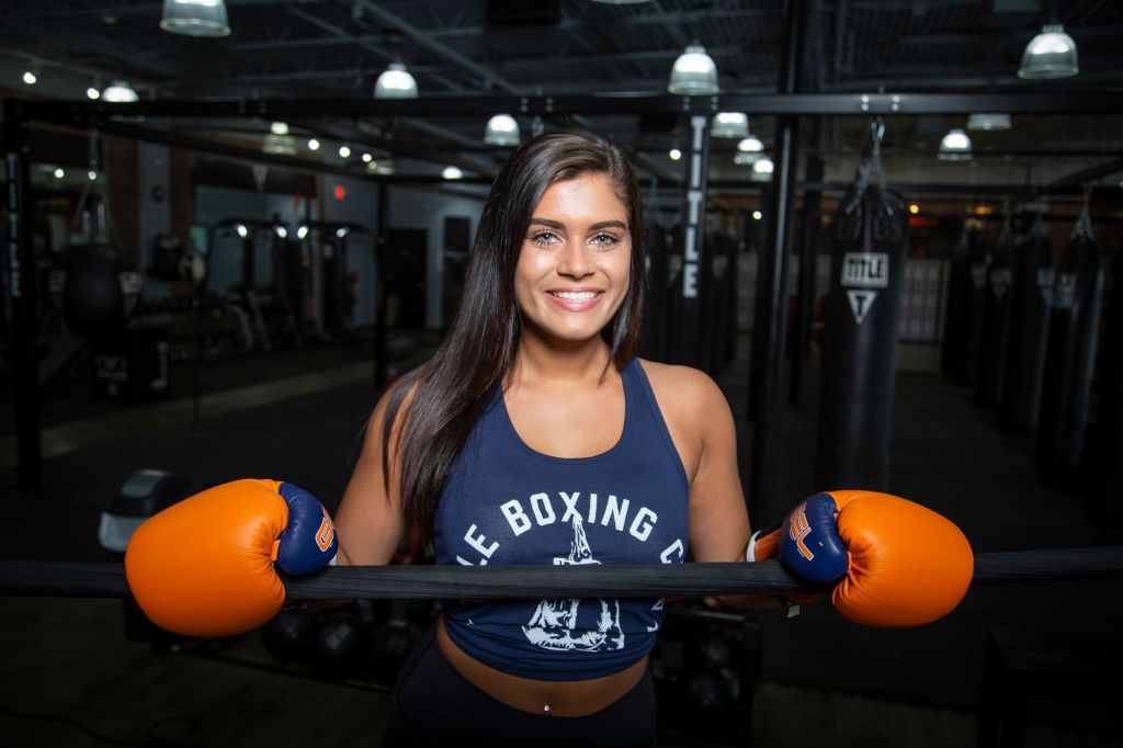 A smiling woman in training gear leaning on the ropes of a boxing ring in her gym